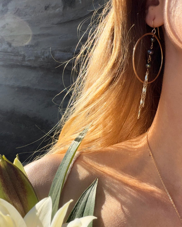Close-up of a person wearing gold hoop earrings with beads, holding white flowers against a blurred background.