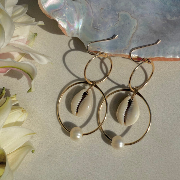 Gold hoop earrings with shell and pearl accents on a textured surface with flowers.