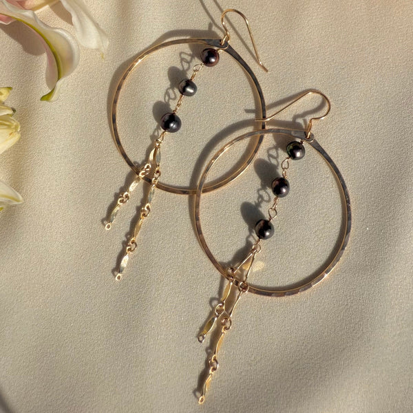 Gold hoop earrings with black pearls on a beige surface with flowers in the background
