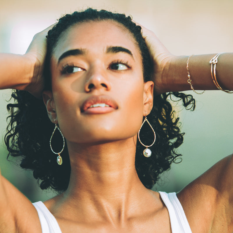 black female model holding her hands on her head, wearing gold hoop earrings with freshwater pearls on them and handmade gold bracelets 