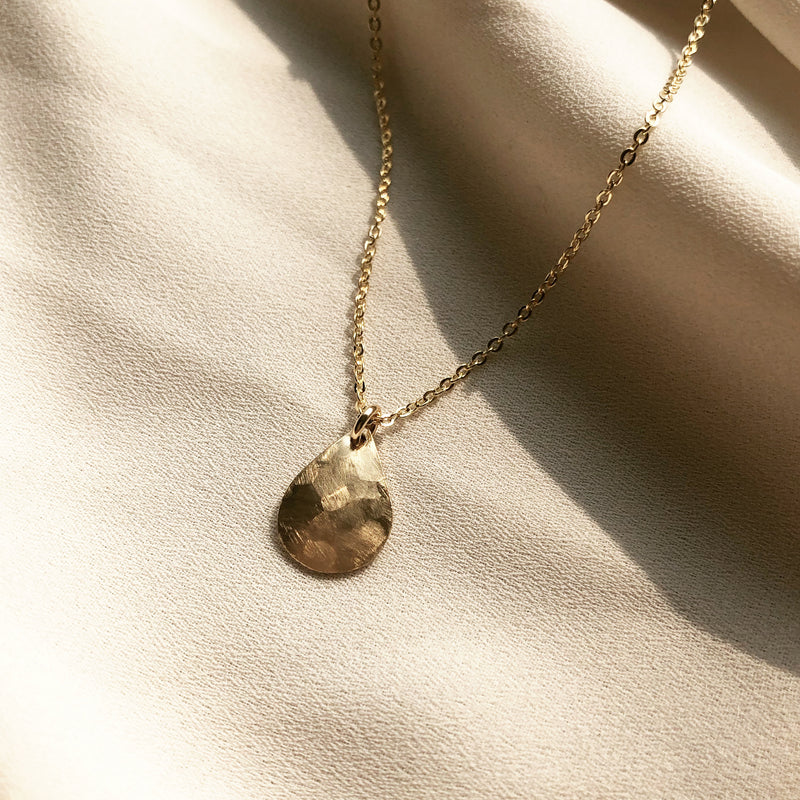 gold water droplet pendant hangs from a gold cable chain, placed on a white fabric 